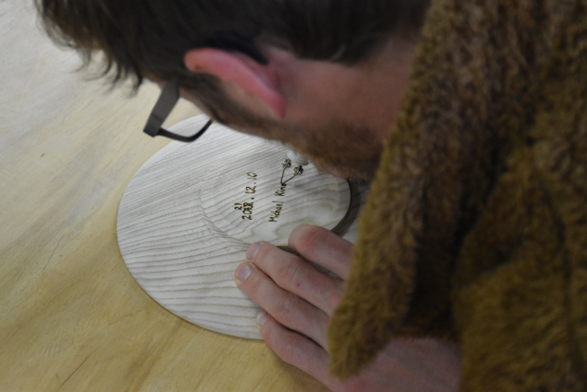 A visitor closely examining and smelling a handcrafted wooden plate with an engraved signature and date, appreciating the natural wood texture.