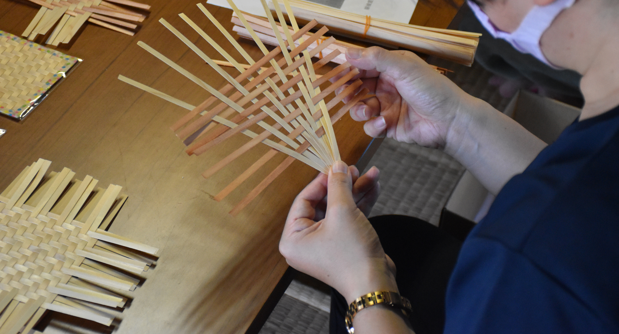 A participant weaving thin bamboo strips into an intricate pattern during a traditional handicraft workshop. Various woven pieces are displayed on the table.