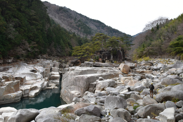 A vast rocky river gorge with a clear blue stream flowing between massive boulders. A hiker explores the rugged landscape, with mountains and dense forests in the background.