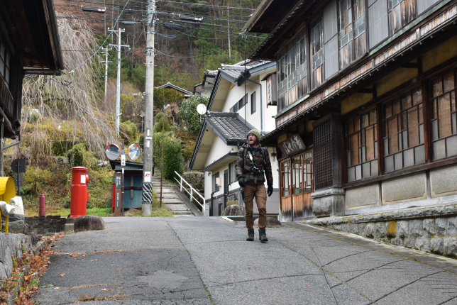 A traveler standing in the middle of a quiet, sloping street in a traditional Japanese village. The street is lined with old wooden houses, and a red postbox adds a nostalgic touch.