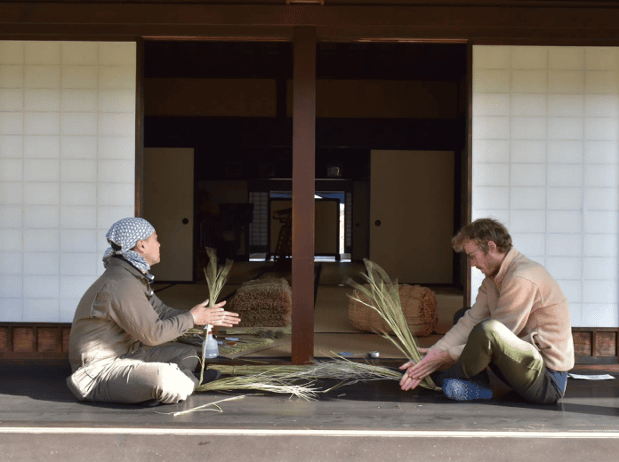 Two people sitting on a wooden veranda, learning traditional straw craft together in a rural Japanese home